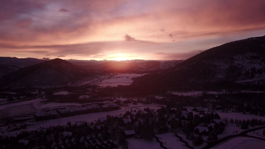Push in style, gorgeous early morning, snowy landscape drone shot in Park City Utah. The sky shines pink purple and orange as the sun peeks over the mountains on the horizon.