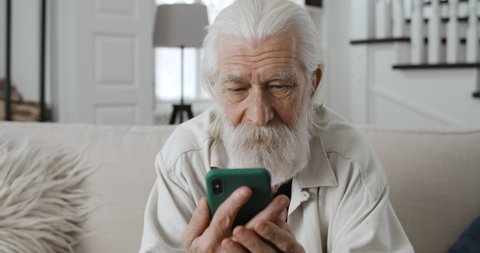 Close up view of old grey haired man chatting with family while looking at phone sreen.Cheerful bearded male retiree reading message and smiling whilesitting on sofa at home.