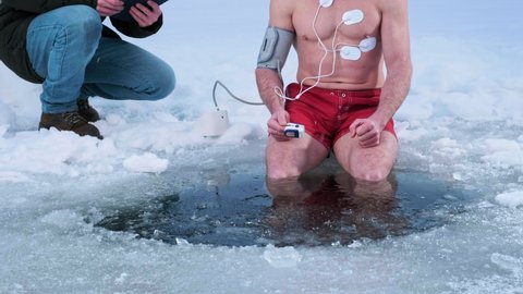 Medical test in the cold. Man sits on the ice with legs immersed into icy water. Oximeter, blood pressure monitor and cardio electrodes are connected to his body, another man comes and checks pressure