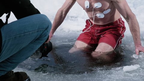 Medical test in the cold. Man films the participant of the medical test. Young man immerses into the icy water with medical electrodes connected to his body