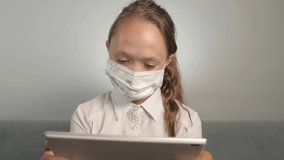 A child in mask plays on his tablet in the room on couch. A girl in a protective mask during an online lesson. A student in medical mask teaches homework at home with tablet. Quarantine, viral danger