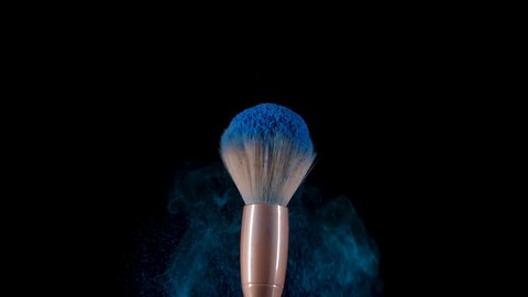 Fluffy makeup brush flinches and creates an explosion of small particles of blue powder against black background. Small particles of shadows fly into space. Great makeup brushes close up. Slow motion.