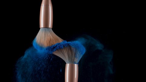 Two makeup brushes collide and cause a swirl of blue powder particles against a black background. Fine dust of crushed colorful paint flies beautifully into the air. Close up. Slow motion.