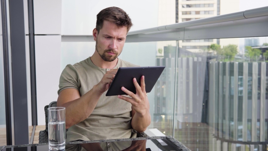 A young handsome man works on a tablet as he sits on a balcony in a luxurious apartment
