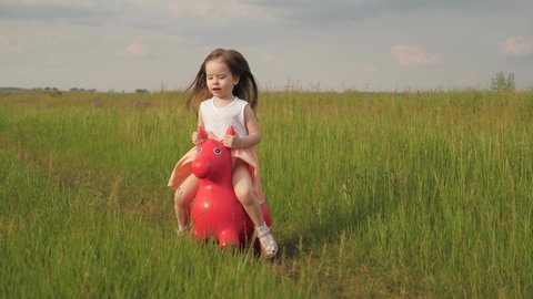 Happy little girl is playing in park and jumping on an inflatable toy donkey. Kid plays in field with his favorite toy horse. Little girl, daughter playing in field outdoors. Family and childhood.