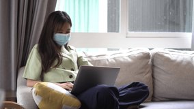 Young woman in medical mask sitting on the sofa in her own house is video call conference with laptop computer raise hand saying hi team at home office, work from home.