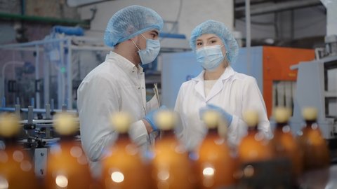 A middle-aged woman at work, in a white coat, disposable cap, medical mask and latex gloves, communicates with her younger colleague next to a conveyor along which plastic bottles filled with a drink
