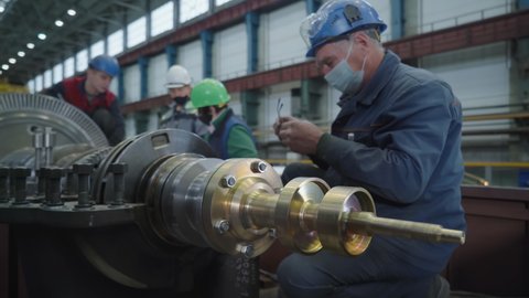 Turbine manufacturer plant. The four workers in protective helmets are getting ready for continuing the process of production of their designated turbine part. Work plans are discussed.