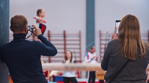 Two proud parents filming their child at the gymnastic tournament on the phone and camera