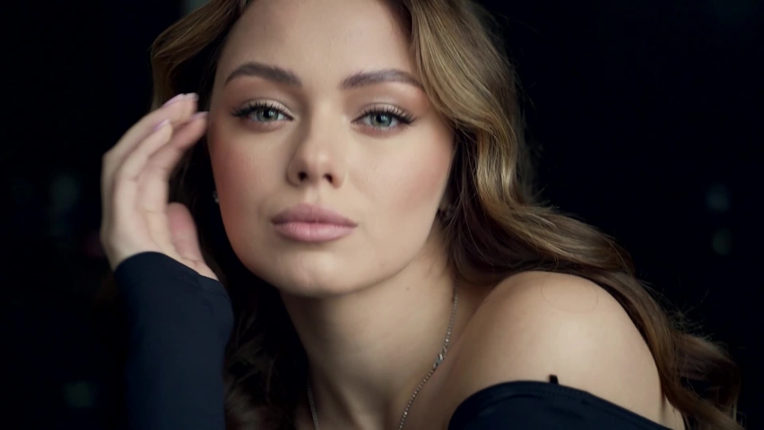 Close-up portrait of a young luxurious sexy woman looking at the camera on a dark background indoors. Face of professional fashion model with make-up miss with blue eyes. Film grain