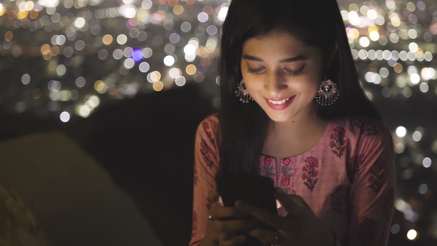 close up shot of a cheerful and attractive young Indian female or a woman sitting on a top and using a mobile phone to browse or to type a text message against the illuminated urban cityscape at night Royalty-Free Stock Footage #1067505860