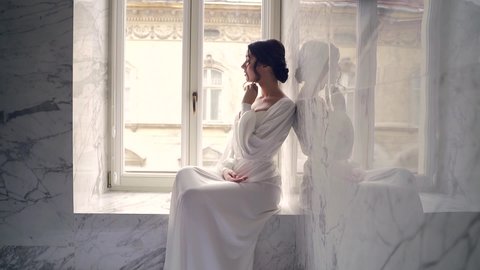 elegant bride in the morning in a dressing gown. young attractive woman in white lingerie in a luxury bathroom interior sitting on the windowsill near the window. looking seductively into the camera