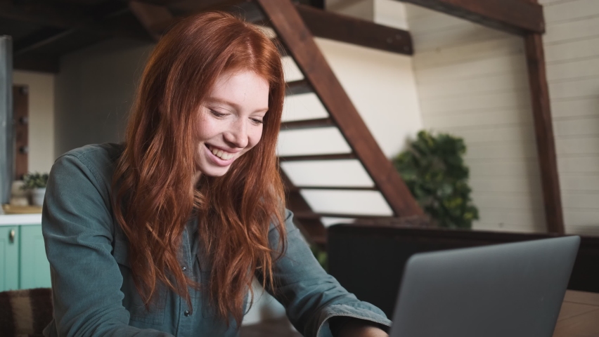 A smiling young redhead woman is using her laptop computer sitting at the table in the apartments
