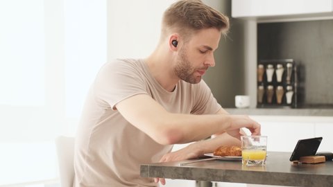 A concentrated young man wearing earbuds is eating breakfast in the kitchen at home