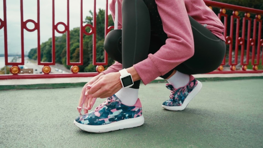 Cropped view of sportswoman with smartwatch tying laces of sneaker on bridge