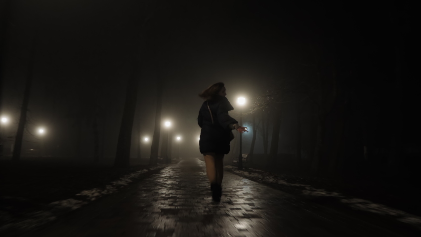 Scared young woman looking back runs away from someone at night in city park