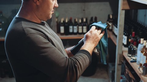 Millennial adult winemaker holding bottle with wine cleaning off dust. Wine factory employee working in cellar warehouse.