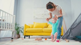 mother holding hands and helping infant son walking on blanket near toys