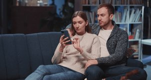 Beautiful young family couple sitting on sofa using mobile phone chatting in social media watching online videos for home entertainment. Technology. Relationships.