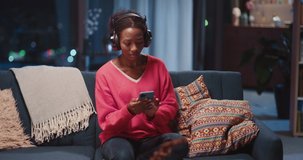 Excited african young woman listening to music in headphones using mobile phone watching clips videos on social media having fun alone at home.