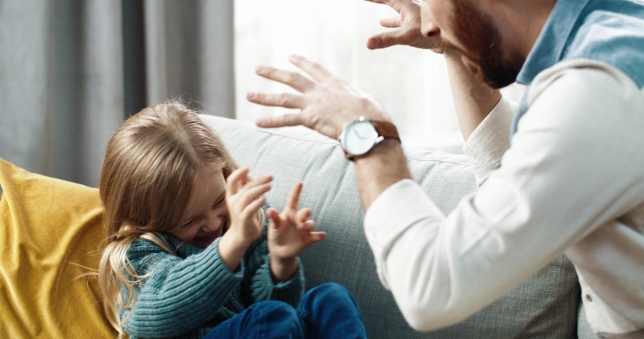 Caring father plays with little daughter sitting at home on couch and tickles her. Cute child is having fun and laughing out loud.