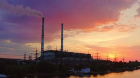Factory chimneys smoke on the river bank in the evening. Beautiful sunset sky reflects in water. Video from drone.