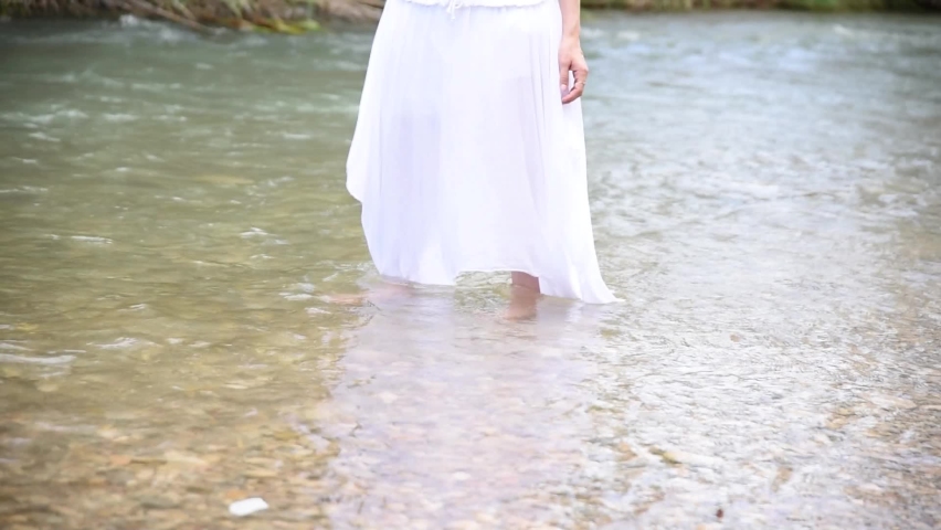 A woman in a white dress in a river. A woman with her feet in the water. A woman stands in a river