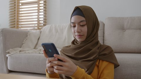 Muslim girl wear hijab using texting smartphone or chat, Asian women happy smile while sitting typing message on mobile in the living room at home morning.