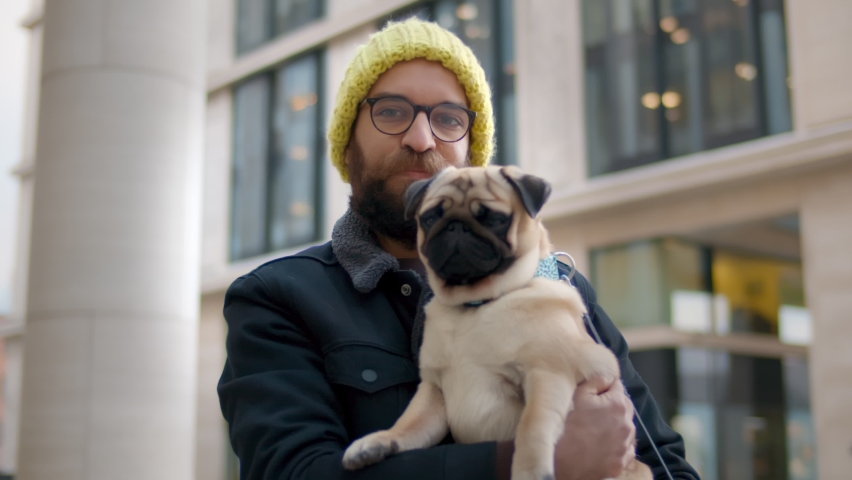 Portrait of young man looking at camera while holding dog on street outdoors. Bearded hipster guy in yellow knitted hat and eyeglasses with pug dog looking at camera near modern building