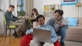 African woman sitting on bean bag and watching funny video with korean friend on laptop while roommates studying at table on background. Students in sharing apartment doing homework and relaxing