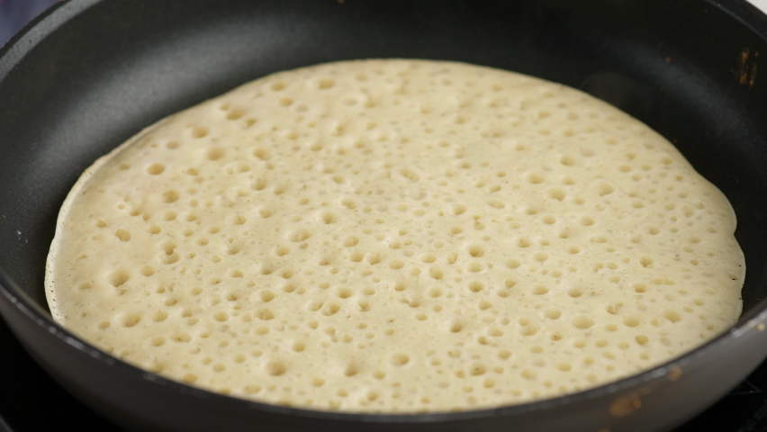 Turning a pancake in a pan. Big round pancake on induction frying pan. Cooking smoke goes up from pancake. Pancakes with kefir. Pancakes with holes. Pancake week. Russian tradition. Royalty-Free Stock Footage #1067558606