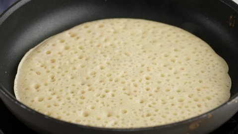 Turning a pancake in a pan. Big round pancake on induction frying pan. Cooking smoke goes up from pancake. Pancakes with kefir. Pancakes with holes. Pancake week. Russian tradition.