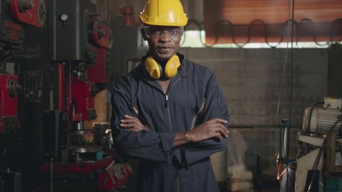 Portrait American industrial black young worker man smiling with yellow helmet and ear protection in front of machine, Happy engineer standing arms crossed at work in industry factory. slow motion
