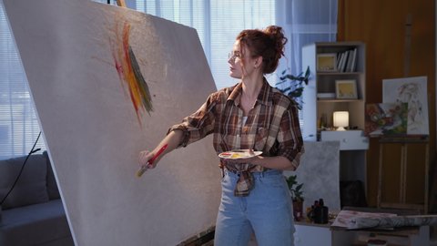 art workshop, girl in glasses draws with brush on large canvas using bright colors, artist uses an easel during modern creativity in slow motion