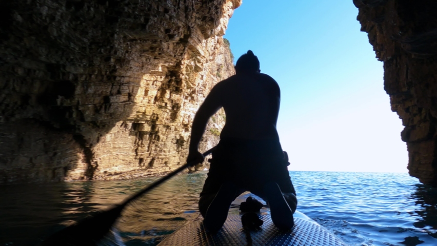 Silhouette of sportsman rowing SUP-board with paddle on sea water through shadow tunnel between old steep cliffs backside view