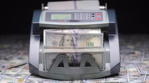 Currency counting machine counts 100 dollar bills or USD banknotes. Cash bill counter money machine stands on scattered dollars. Automatic mechanism for bank financial operations. Finance concept.
