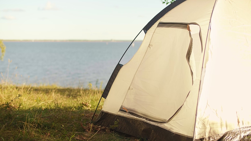 A young tourist in a cap comes to his tent standing on the seashore, river, lake, closes it and goes to the campground. Tourist weekend on the shore of sea, lake, river.