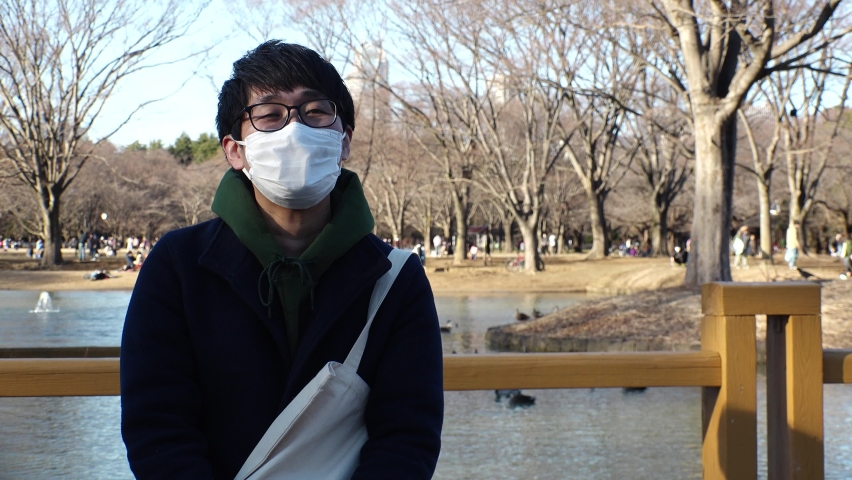 Asian (Japanese) young man wearing white surgical mask sitting on the bench at the park in winter daytime. One person, late twenties, black hair with glasses. Coronavirus (COVID-19) concept shot.
