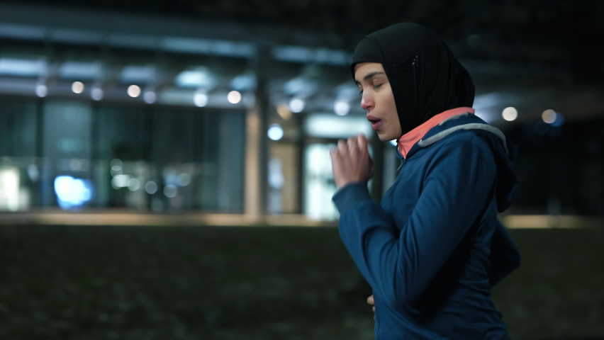 Cinematic shot of young arab sportswoman with athletic body in hijab running with effort and dedication in city center with snow falling at night. Concept:determination,woman power,goal achievement. Royalty-Free Stock Footage #1067572343