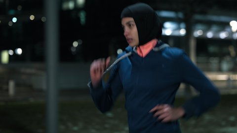 Cinematic shot of young arab sportswoman with athletic body in hijab running with effort and dedication in city center with snow falling at night. Concept:determination,woman power,goal achievement.