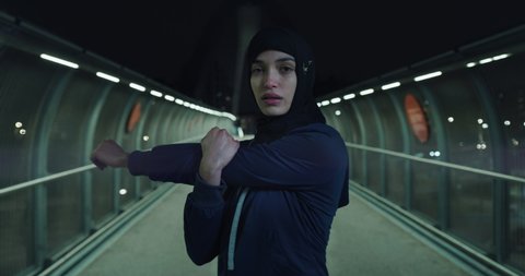 Cinematic shot of young arab sportswoman in hijab is warming up and looking in camera before run with dedication in city center at night. Concept of determination, woman power and goal achievement.