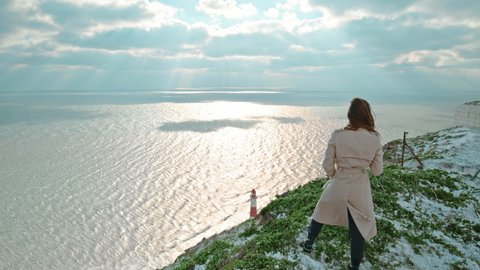 A young girl watching the ocean at the edge on top of a rocky seashore in 4K. A woman smartly dressed enjoying a lighthouse view during winter on a windy day, the wind blowing hair in slow motion.