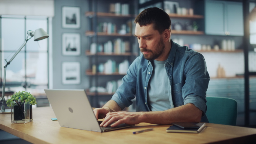 Handsome Caucasian Man Working on Laptop Computer while Sitting on a Sofa Couch in Stylish Cozy Living Room. Freelancer Working From Home. Browsing Internet, Using Social Networks, Being Serious.