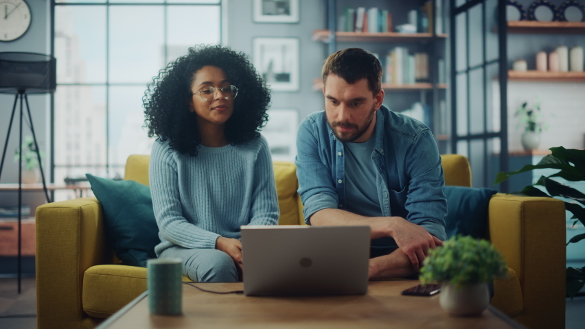 Diverse Multiethnic Couple are Sitting on a Couch Sofa in Stylish Living Room and Choosing Items to Buy Online with Laptop Computer. Friends or Colleagues are Discuss Work Projects and Give High Five. Royalty-Free Stock Footage #1067577206
