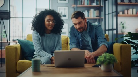 Diverse Multiethnic Couple are Sitting on a Couch Sofa in Stylish Living Room and Choosing Items to Buy Online with Laptop Computer. Friends or Colleagues are Discuss Work Projects and Give High Five.