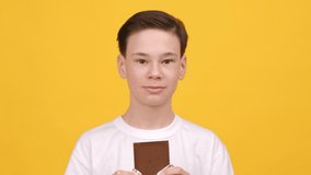 Sweet Tooth. Happy Teen Boy Eating Chocolate Bar Enjoying Tasty Snack Posing Standing On Yellow Studio Background, Looking At Camera. Sugary Food, Unhealthy Dessert Concept