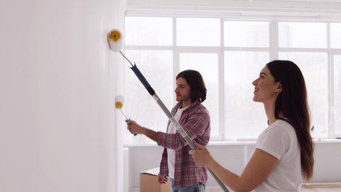 Young married couple making repair together, painting white wall in empty apartment with rollers, slow motion