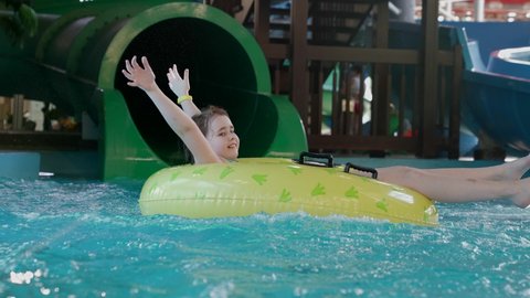 A little girl slips out of the pipe of a green water slide in a water park, she plops into the pool on an inflatable circle.