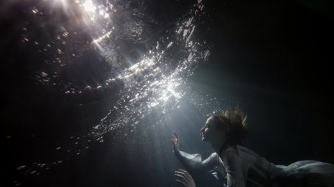 A beautiful woman in a flowing dress floats under the water on a dark background, she swims to the surface of the water and comes up, creating a lot of bubbles.