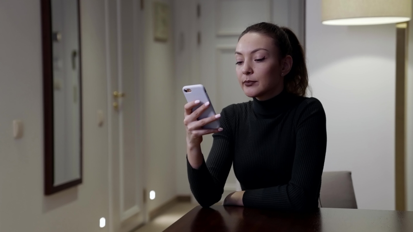 A brooding brunette woman sits at a table in the room and looks at her smartphone, pursing her lips, calling someone while holding the phone to her ear.